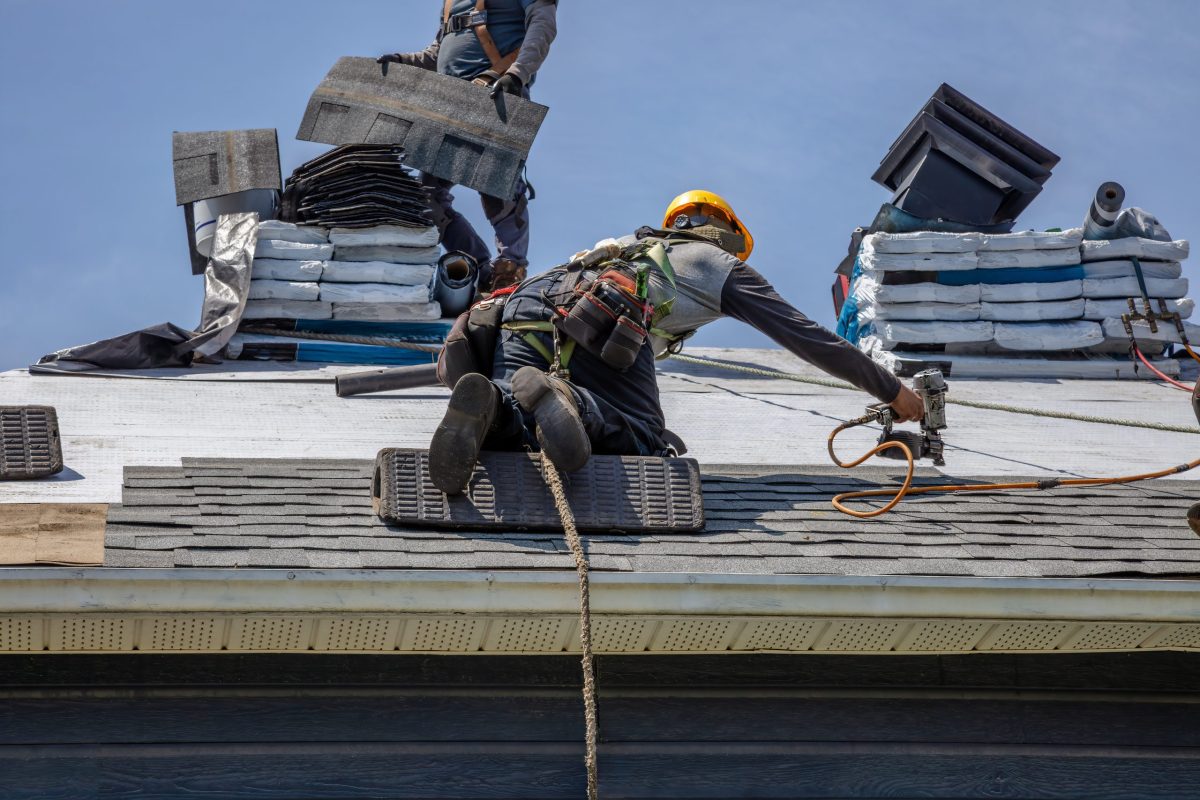 Professional workers using pneumatic nailer and safety helmets applying roofing materials on home exterior