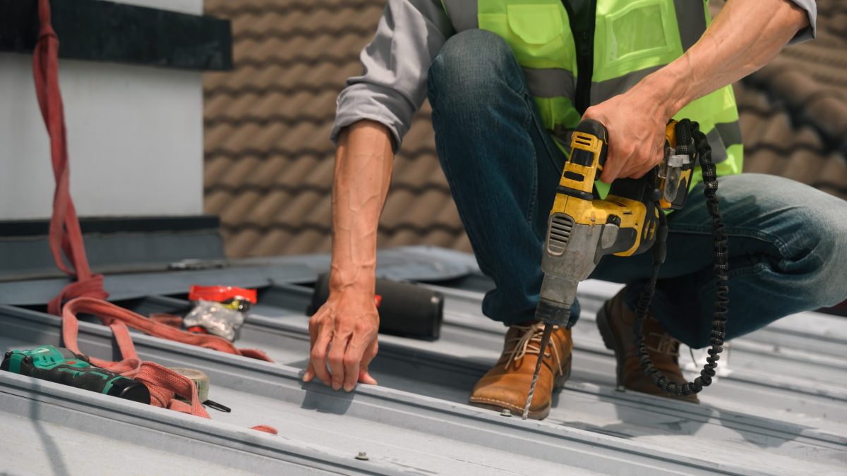 Close up of a professional roofer in a safety vest drills into a metal roof panel during maintenance work, with tools and harness visible nearby.